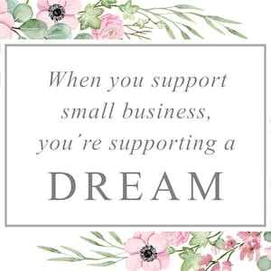May include: A white rectangular sign with the text "When you support small business, you're supporting a DREAM" in gray. The sign is framed with pink flowers, green leaves, and blue accents.