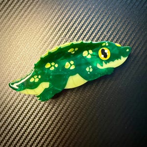 May include: A green and yellow alligator-shaped brooch. The brooch has a glossy finish with a marbled effect. The alligator's body is green with yellow spots, and the underside is yellow. The eyes are yellow with black pupils. The brooch is a unique accessory.