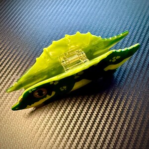May include: A green and yellow toy alligator with a clear, internal mechanism. The alligator has a detailed design with yellow and green spots and a spring-like component inside. The toy is set against a textured, dark background.