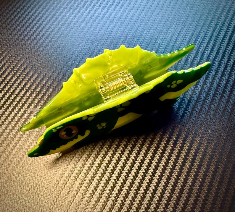 May include: A green and yellow toy alligator with a clear, internal mechanism. The alligator has a detailed design with yellow and green spots and a spring-like component inside. The toy is set against a textured, dark background.