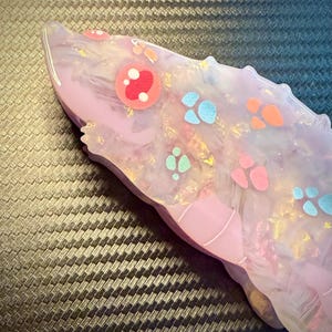 May include: A light purple, resin-cast object with a rounded shape, decorated with colorful paw prints and circular designs. The object has a glossy finish and a textured appearance, with hints of gold and white throughout.