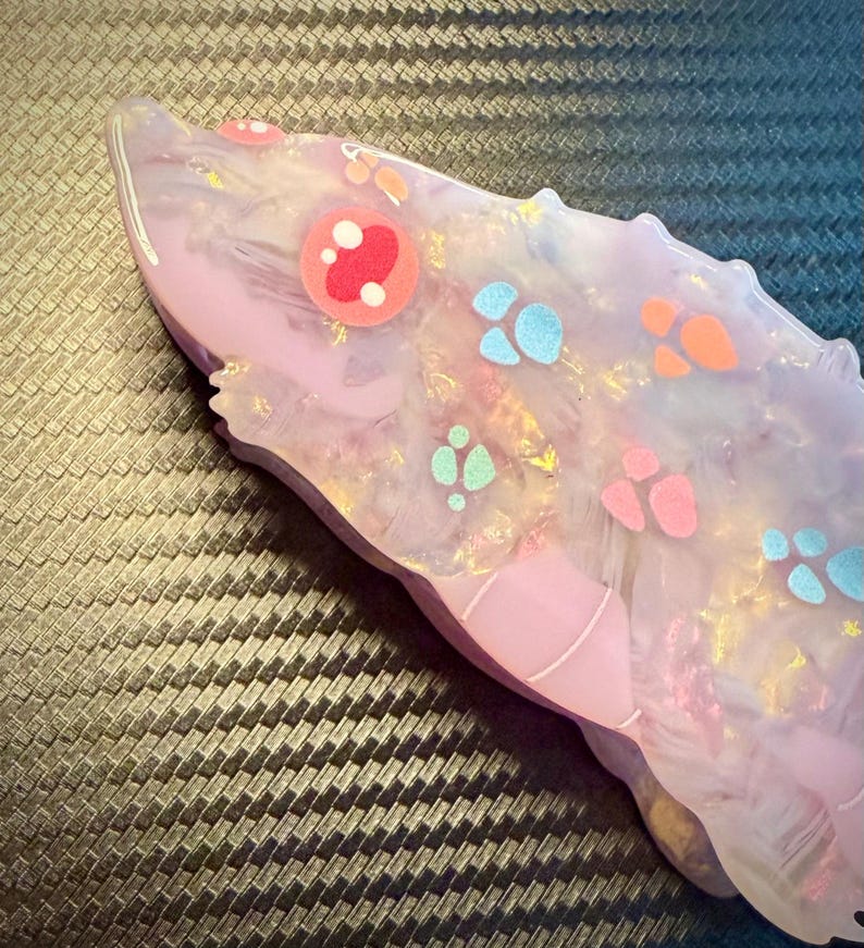 May include: A light purple, resin-cast object with a rounded shape, decorated with colorful paw prints and circular designs. The object has a glossy finish and a textured appearance, with hints of gold and white throughout.