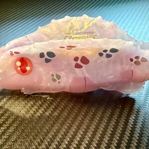 May include: A unique, abstract sculpture in shades of pink and white, adorned with colorful, patterned accents. The piece features a textured, organic form with a small, gold-colored detail on top. The sculpture is set against a dark, patterned surface.
