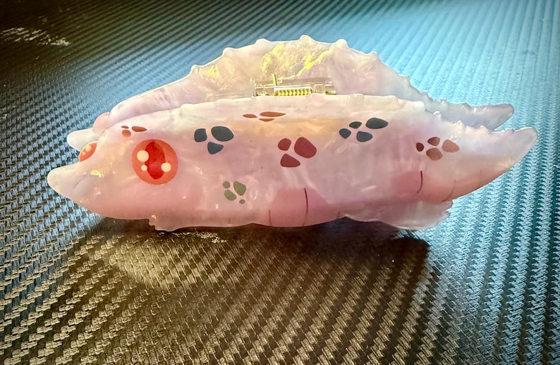 May include: A unique, abstract sculpture in shades of pink and white, adorned with colorful, patterned accents. The piece features a textured, organic form with a small, gold-colored detail on top. The sculpture is set against a dark, patterned surface.