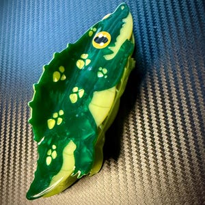 May include: A handcrafted, green and yellow alligator figurine. The figurine features a detailed design with yellow spots and a yellow eye with a black pupil. The alligator's body is a vibrant green color, with a glossy finish.