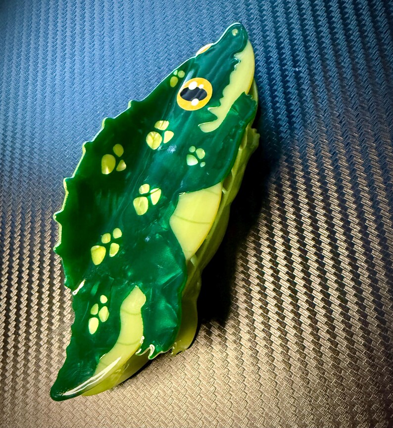 May include: A handcrafted, green and yellow alligator figurine. The figurine features a detailed design with yellow spots and a yellow eye with a black pupil. The alligator's body is a vibrant green color, with a glossy finish.
