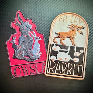 Puede incluir: Dos parches bordados, uno rojo con conejos negros y el texto "OWSLA" y otro beige con un conejo marrón y el texto "CHIEF RABBIT".