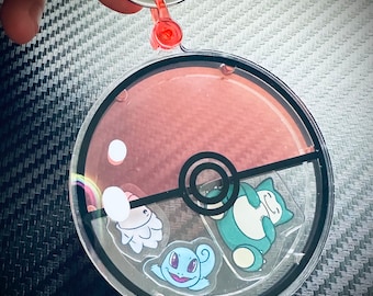 May include: A clear plastic Pokeball keychain with a red and black design. The keychain features three Pokemon characters: Squirtle, Bulbasaur, and Snorlax.