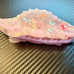 May include: A pink and translucent hair clip with a spiky design, adorned with colorful accents. The clip features a series of small, decorative shapes in shades of orange, blue, and pink. The clip is approximately 4 inches long.