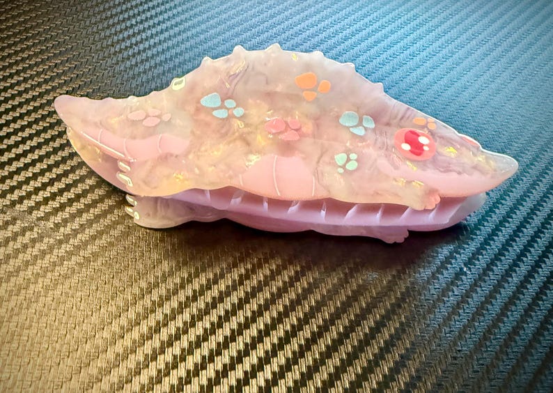 May include: A pink and translucent hair clip with a spiky design, adorned with colorful accents. The clip features a series of small, decorative shapes in shades of orange, blue, and pink. The clip is approximately 4 inches long.