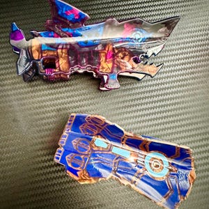 May include: Two blue and gold resin-cast figurines. The first is a shark-shaped weapon with a purple and gold design. The second is a hand-shaped weapon with a blue and gold design.