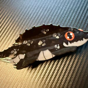 May include: A black and white alligator-shaped decorative object with red eyes and white teeth. The alligator has a textured, glossy finish with silver accents. The object is likely a decorative figurine or trinket.