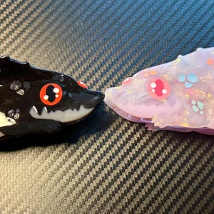 May include: Two resin alligator-like figurines. One is black with white and gray accents, red eyes, and a toothy grin. The other is a translucent lavender with pink, blue, and orange details and red eyes. Both have a textured, spiky back.