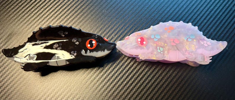 May include: Two resin alligator-like figurines. One is black with white and gray accents, red eyes, and a toothy grin. The other is a translucent lavender with pink, blue, and orange details and red eyes. Both have a textured, spiky back.