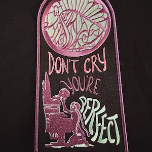Può includere: Una toppa ricamata nera con cuciture rosa e verdi. La toppa presenta un'immagine stilizzata di una persona seduta su un trono con il testo "Don't cry you're perfect" in rosa e verde.