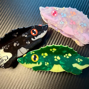 May include: Three decorative snake-shaped hair clips in the image. One is black with white accents and orange eyes, another is pink with colorful spots, and the third is green with yellow accents. The clips are made of a glossy material.