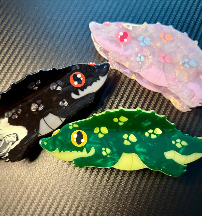 May include: Three decorative snake-shaped hair clips in the image. One is black with white accents and orange eyes, another is pink with colorful spots, and the third is green with yellow accents. The clips are made of a glossy material.