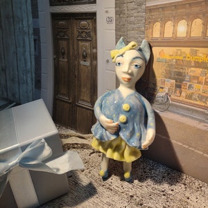 Op de afbeelding: Een blauw en geel keramiek beeldje van een fantasievol personage met een strik, staand voor een gebouw met een deur en een bord met de tekst "Winkel Dumple".