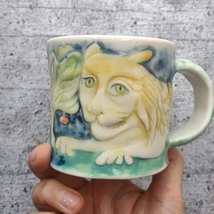 Peut inclure: Une tasse en céramique avec un motif peint à la main d'un chat jaune aux yeux verts sur un fond bleu. Le chat est représenté avec une patte levée et une baie suspendue à une feuille verte.