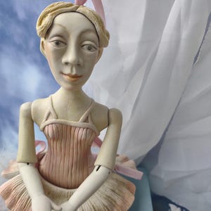 Könnte beinhalten: Eine Porzellan-Ballerina-Puppe mit einem rosa Tutu und einem rosa Band im Haar. Die Puppe sitzt auf einer blauen Oberfläche.