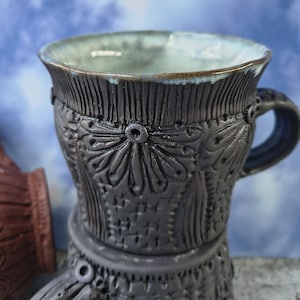 Könnte beinhalten: Eine handgefertigte schwarze Keramiktasse mit detailliertem Blumenmuster und blau-grünem Innenraum. Die Tasse hat einen Henkel und ist etwa 10 cm hoch. Das Design umfasst strukturierte vertikale Linien und dekorative Elemente.
