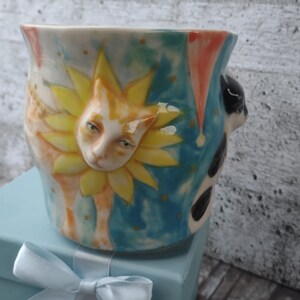 Op de afbeelding: Een keramische mok met een handgeschilderd ontwerp met een kattengezicht met een zonneschijn erachter. De mok is versierd met gele, oranje, blauwe en witte verf.