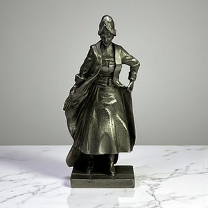 May include: A detailed, antique-style metal sculpture of a woman in a long coat and dress, holding the coat open. The sculpture is dark gray and stands on a rectangular base. The woman's hair is styled in a bun.