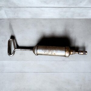 May include: A vintage, silver-toned grease gun with a handle and nozzle. The cylindrical body has text printed on it. The tool is designed for applying grease to machinery and equipment. The handle is a loop shape.