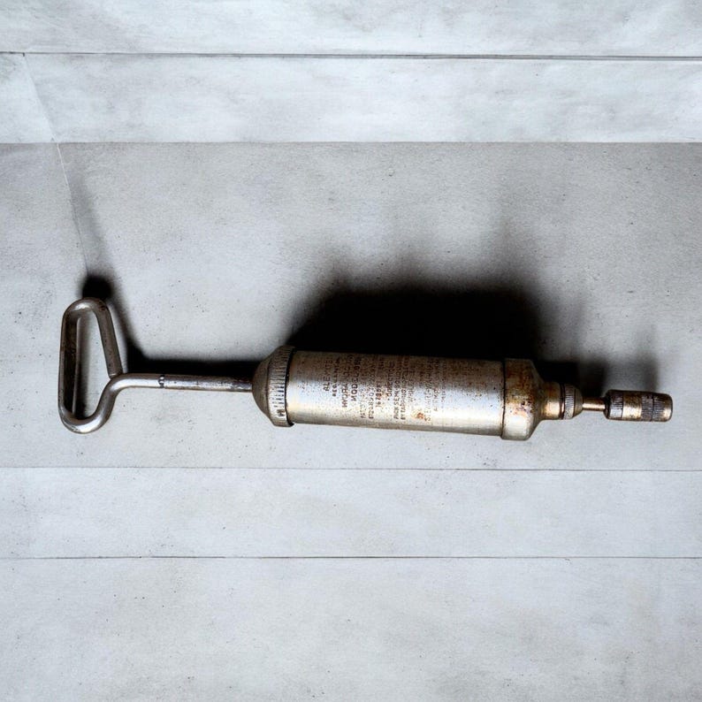 May include: A vintage, silver-toned grease gun with a handle and nozzle. The cylindrical body has text printed on it. The tool is designed for applying grease to machinery and equipment. The handle is a loop shape.