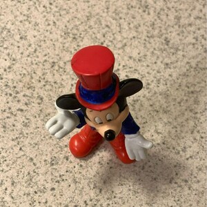 Pu&ograve; includere: Una statuetta di Topolino con un cappello a cilindro rosso con una fascia blu, una giacca blu, pantaloni rossi e guanti bianchi. Il personaggio dei cartoni animati classico ha naso e orecchie neri ed &egrave; in posa con le braccia tese.