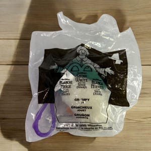 May include: A clear plastic bag with a purple handle contains a small plastic toy of Grumpy from the Disney movie Snow White and the Seven Dwarfs. The bag has text in English, French, and Spanish.