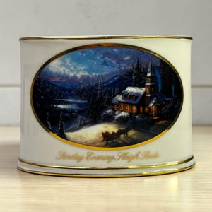May include: A white ceramic cylinder with a gold rim and accents. An oval image depicts a winter scene with a church, horse-drawn sleigh, and text that reads "Sunding Evening Sleigh Ride".