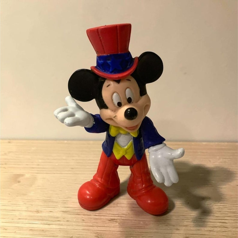Pu&ograve; includere: Una statuina di Topolino in costume da mago. La figura indossa un cappello a cilindro rosso con finiture blu, una giacca blu, pantaloni rossi e guanti bianchi. La statuina &egrave; su una superficie di legno.