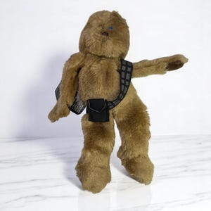 May include: A brown plush Chewbacca toy with a black bandolier and a small black pouch. The toy has blue eyes and is standing on a white marble surface.