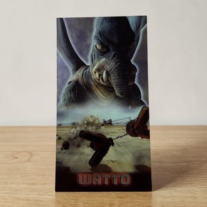 May include: A rectangular card featuring a character with large eyes, a trunk-like nose, and large ears. The card has a desert scene with flying vehicles and the name "WATTO" at the bottom.
