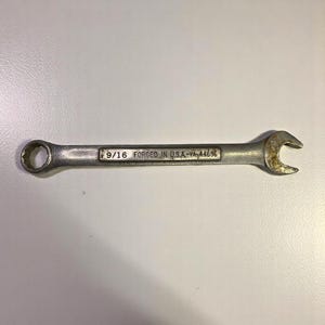 May include: A silver wrench with a 9/16 inch opening on one end and a curved opening on the other. The text "9/16 FORGED IN U.S.A.-VA-44696" is printed on the wrench.