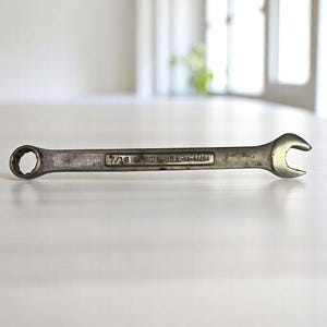 May include: A silver wrench with a closed end and an open end. The wrench has the text "7/16 FORGED IN U.S.A.-VA-44694" printed on it. The tool is designed for tightening or loosening nuts and bolts.
