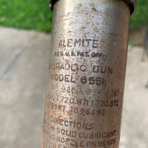 May include: Close-up of a vintage Alemite hydraulic gun, Model 6551, with visible text including "REG. U.S. PAT. OFF." and patent numbers. The metal surface shows signs of age and wear, with rust and discoloration.