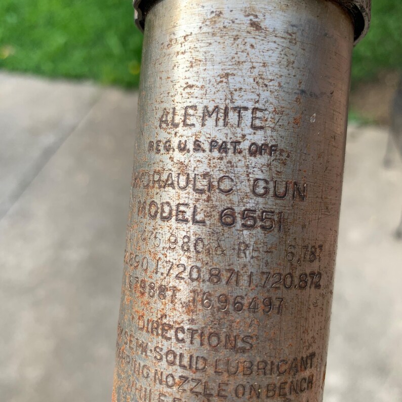 May include: Close-up of a vintage Alemite hydraulic gun, Model 6551, with visible text including "REG. U.S. PAT. OFF." and patent numbers. The metal surface shows signs of age and wear, with rust and discoloration.