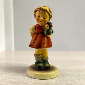May include: A vintage ceramic figurine of a young girl in a red dress, holding a doll. The figurine has a yellow base, yellow hair, and green shoes. The girl's cheeks are rosy, and her eyes are closed. The doll is wearing a green jacket.