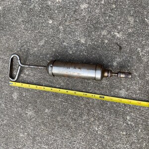 May include: A vintage, metal grease gun with a handle and nozzle. The gun is silver with some rust and is approximately 12 inches long. A yellow measuring tape is placed next to the gun.