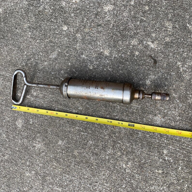 May include: A vintage, metal grease gun with a handle and nozzle. The gun is silver with some rust and is approximately 12 inches long. A yellow measuring tape is placed next to the gun.