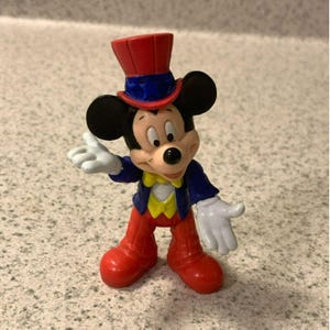 Pu&ograve; includere: Piccola statuina di Topolino in abito da mago. Mickey indossa un cappello a cilindro rosso, una giacca blu, pantaloni rossi e un gilet giallo. Le sue mani guantate bianche sono aperte. Un oggetto da collezione.