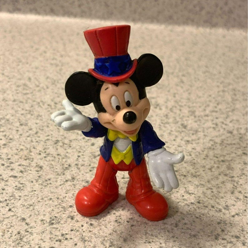 Pu&ograve; includere: Piccola statuina di Topolino in abito da mago. Mickey indossa un cappello a cilindro rosso, una giacca blu, pantaloni rossi e un gilet giallo. Le sue mani guantate bianche sono aperte. Un oggetto da collezione.