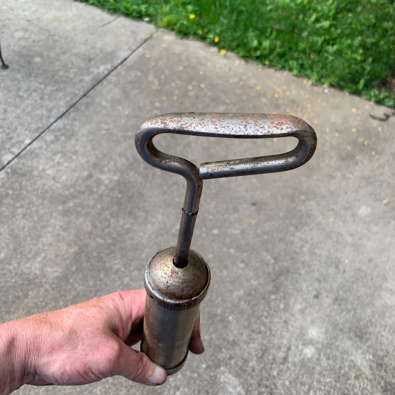May include: An antique grease gun with a cylindrical body and a curved handle. The metal tool shows signs of age with rust and wear, suggesting it's a vintage item. The handle is a loop shape, and the body is a dark, metallic color.