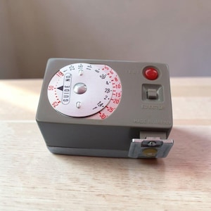 May include: A gray vintage camera light meter with a circular dial featuring numbers and a needle. It has a red button labeled "TEST" and the text "MADE IN JAPAN". A metal bracket is attached to the bottom.