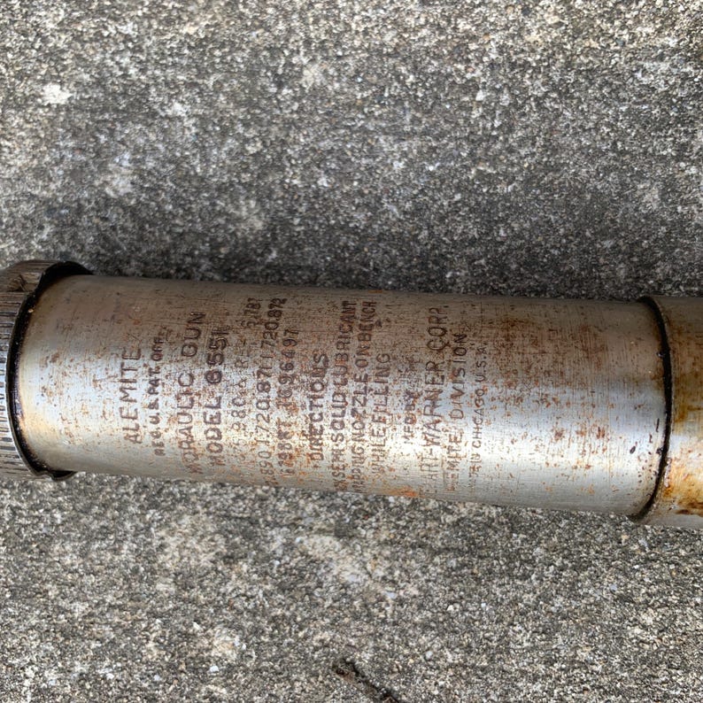 May include: A vintage Alemite Model 655 hydraulic gun, cylindrical in shape, with visible text including "Alemite" and "Hydraulic Gun." The metal body shows signs of age and wear, with a threaded end and a nozzle. The text provides instructions for use.