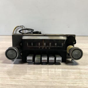 May include: Vintage Ford car radio with a black faceplate, chrome knobs, and push buttons. The radio dial displays numbers from 5 to 16. Wires and a plug extend from the back of the radio. The radio is set on a light wood surface.
