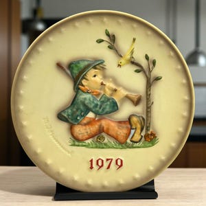 May include: A decorative plate featuring a young boy playing a flute, with a yellow bird perched on a tree branch. The boy wears a green hat and jacket, and orange pants. The plate is cream-colored with the year "1979" in red.