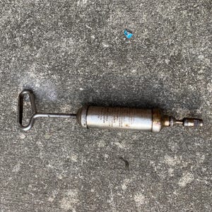 May include: A vintage grease gun with a silver metal body and a handle. The gun has a nozzle on one end and a handle on the other. The body of the gun has text printed on it. The grease gun is designed for applying grease to machinery.